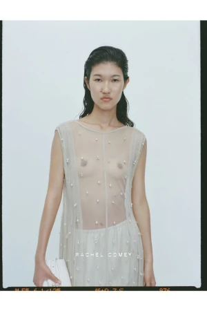 Portrait of an Asian female fashion model wearing a sheer beige dress with bead accents, holding a white clutch against a bright backdrop.