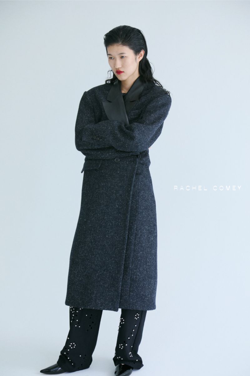 Elegant Asian woman poses in a long, dark coat with arms crossed, paired with distinctive black trousers, set against a stark white backdrop.