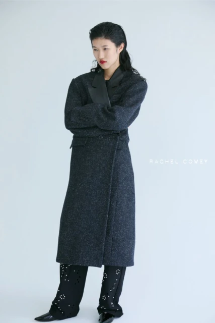 Elegant Asian woman poses in a long, dark coat with arms crossed, paired with distinctive black trousers, set against a stark white backdrop.