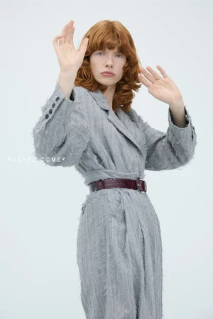 A redhead adult female poses in a grey, fringe-textured jacket dress with a dark belt against a white background in a studio shot.