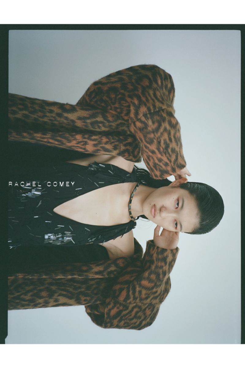 A woman in a black dress and animal print coat poses with her hand near her face in a studio shot, with visible text 'RACHEL COMEY'.
