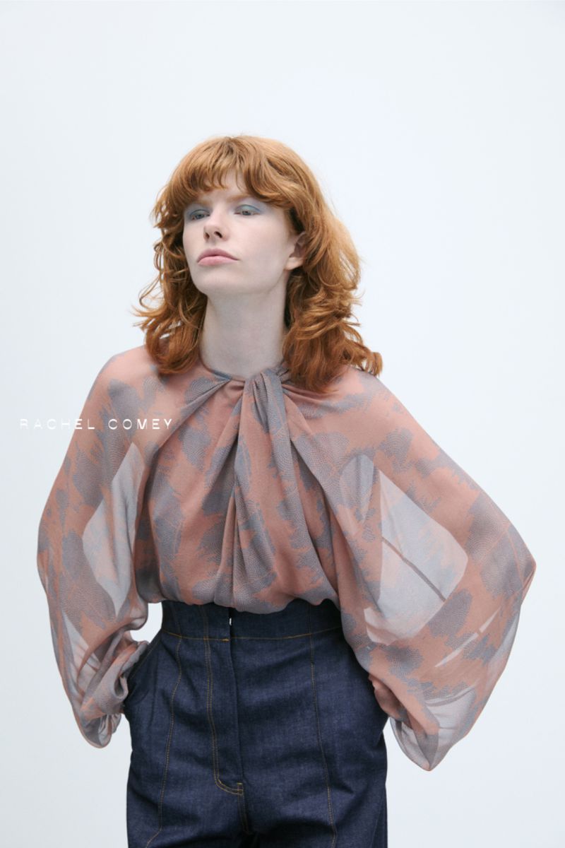 Fashion portrait of a red-haired woman wearing a sheer, patterned top and high-waisted dark denim jeans against a white background.
