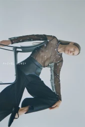 Model wearing a sheer black embellished top and dark trousers seated on a dark chair against a white seamless background.