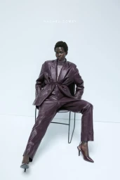 Black model seated on a minimalist chair wearing a dark plum leather suit and heels against a white studio background.