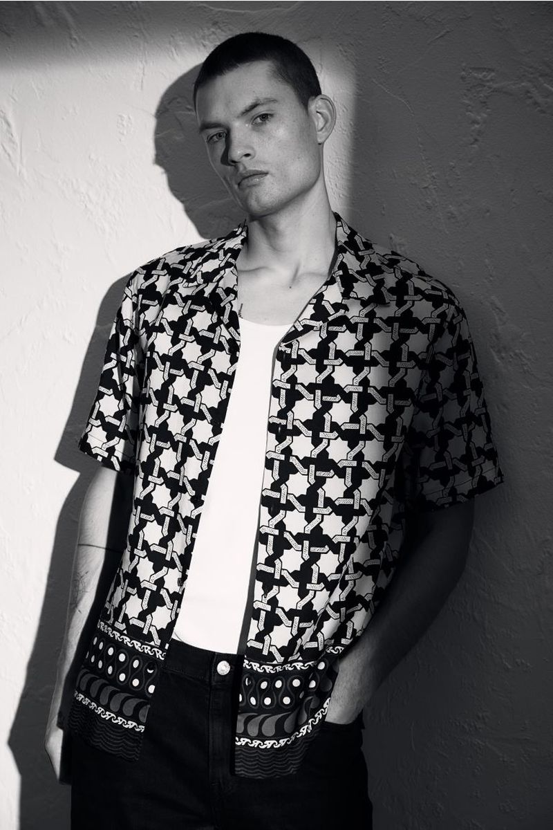 Monochrome portrait of a young man in a patterned shirt, leaning against a textured wall with light and shadow, providing a stark contrast.