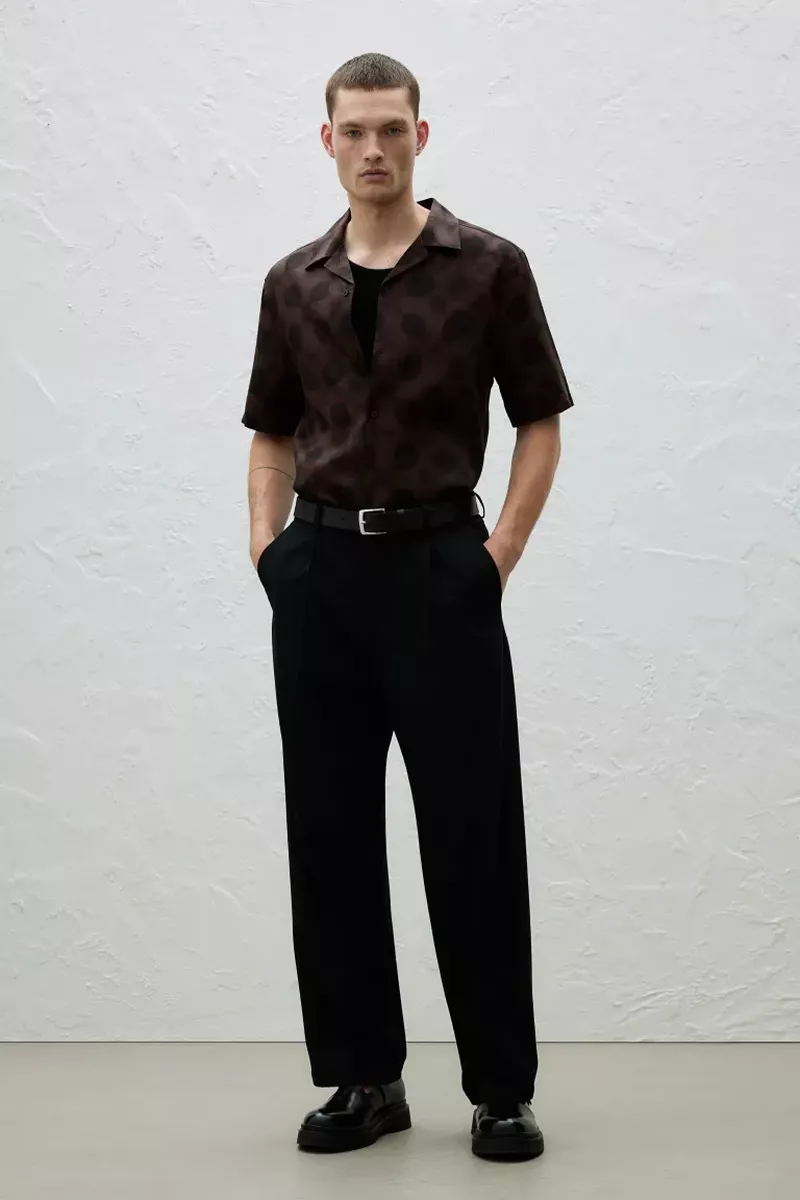 Full-length studio shot of a male model in a patterned shirt, black pants, and shiny black shoes against a textured neutral backdrop.
