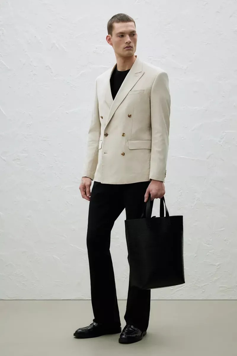 Adult man in a beige double-breasted blazer and black pants, holding a black tote bag, standing against a textured off-white wall.