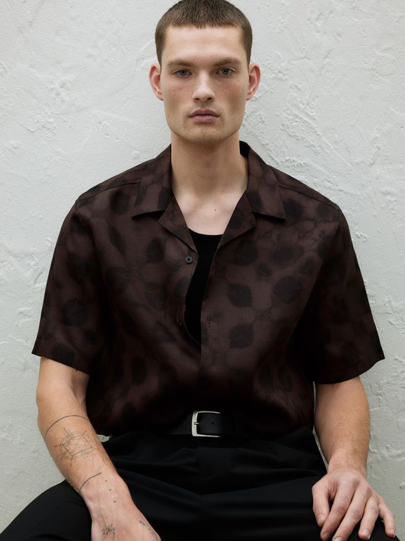 Portrait of a young adult man in a patterned brown shirt and black pants, sitting indoors against a textured neutral background.