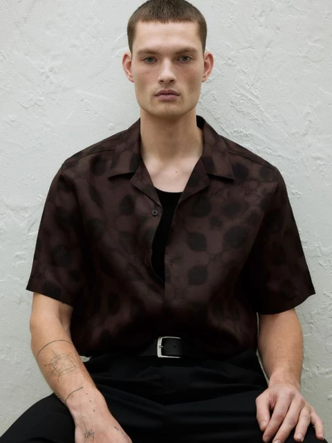 Portrait of a young adult man in a patterned brown shirt and black pants, sitting indoors against a textured neutral background.