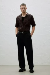 Model wearing a patterned short-sleeve shirt and black trousers against a textured background