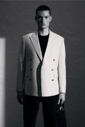 Man wearing a light double-breasted blazer and dark t-shirt against a textured studio background.