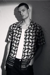 Monochrome portrait of a male model wearing a patterned short-sleeve shirt over a white tank top against a textured wall.
