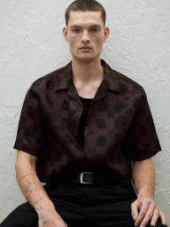 A young male model with a short haircut wearing a patterned brown short-sleeve shirt against a textured white wall