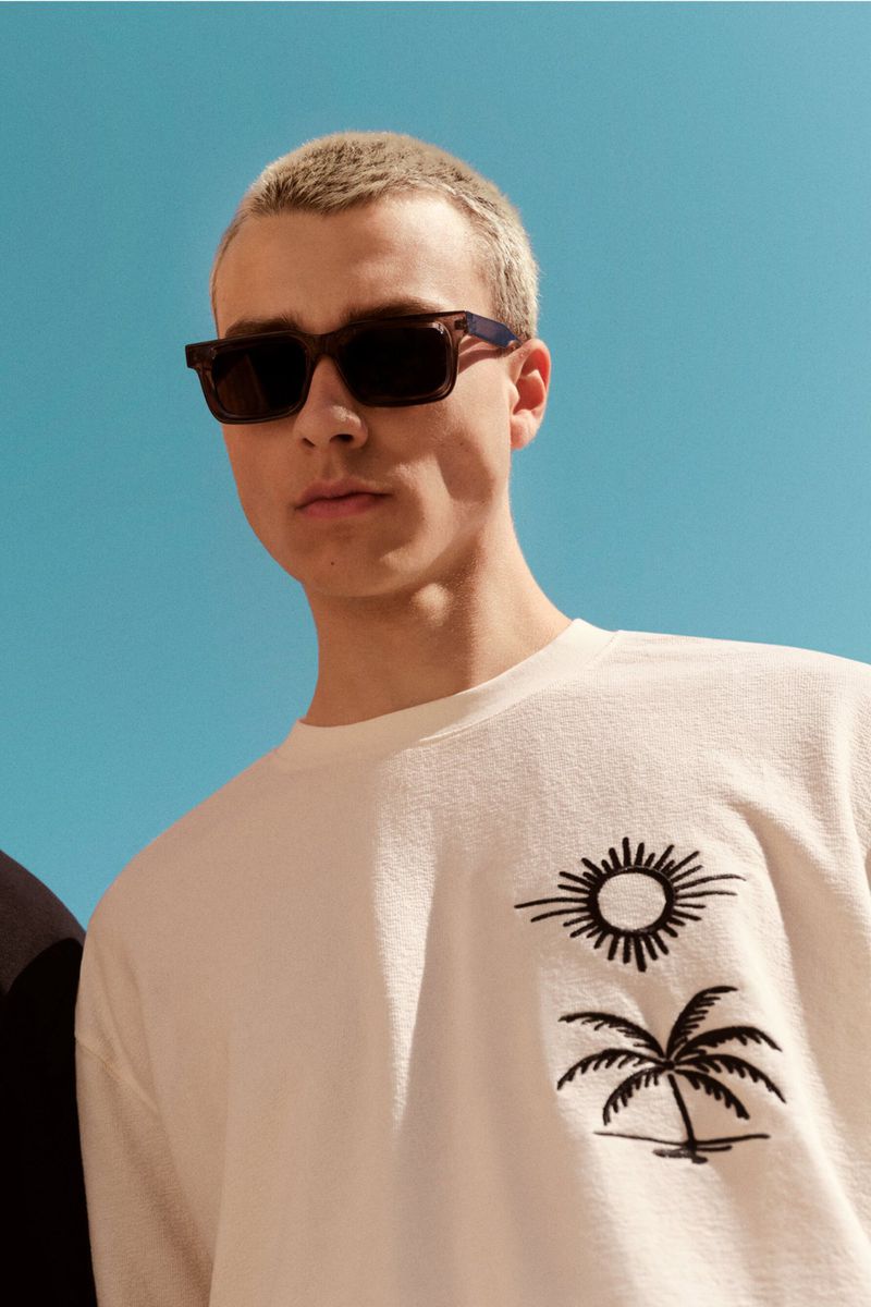 Man in sunglasses and white t-shirt with sun and palm tree design against a blue sky, shot from a low angle.