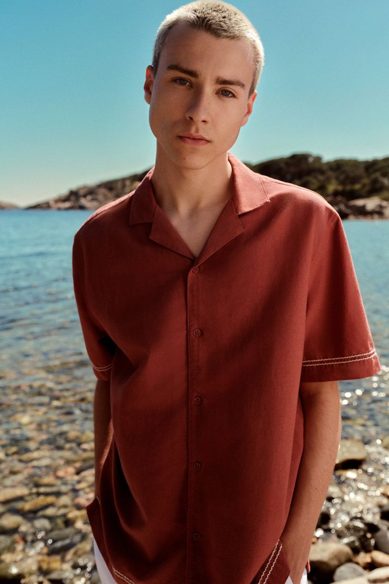 Young adult man with platinum blond hair and casual rust-colored shirt against a coastal backdrop on a sunny day.