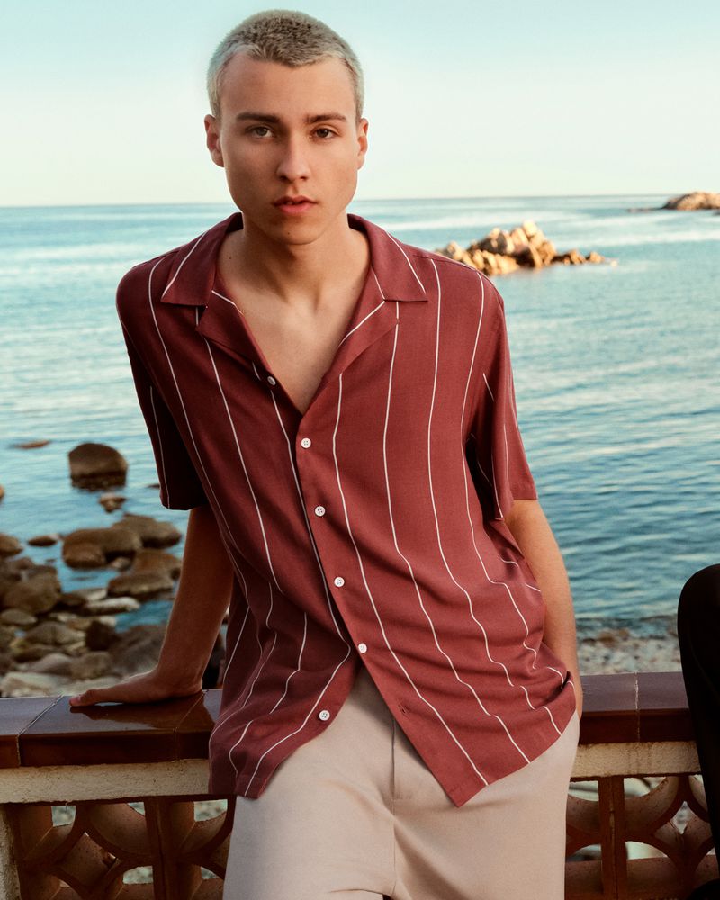 Young man in striped shirt poses on a balcony overlooking the sea. Clear sky, rocks, and coastline provide a serene backdrop.