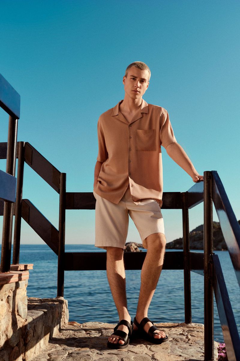 Young male model poses on stone steps overlooking the ocean. He wears a tan shirt, beige shorts, and black sandals, under a bright sky.