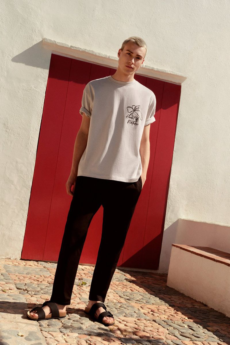 Male fashion model in casual gray t-shirt, black pants, and sandals poses against a red door and whitewashed wall on a stone-paved surface.