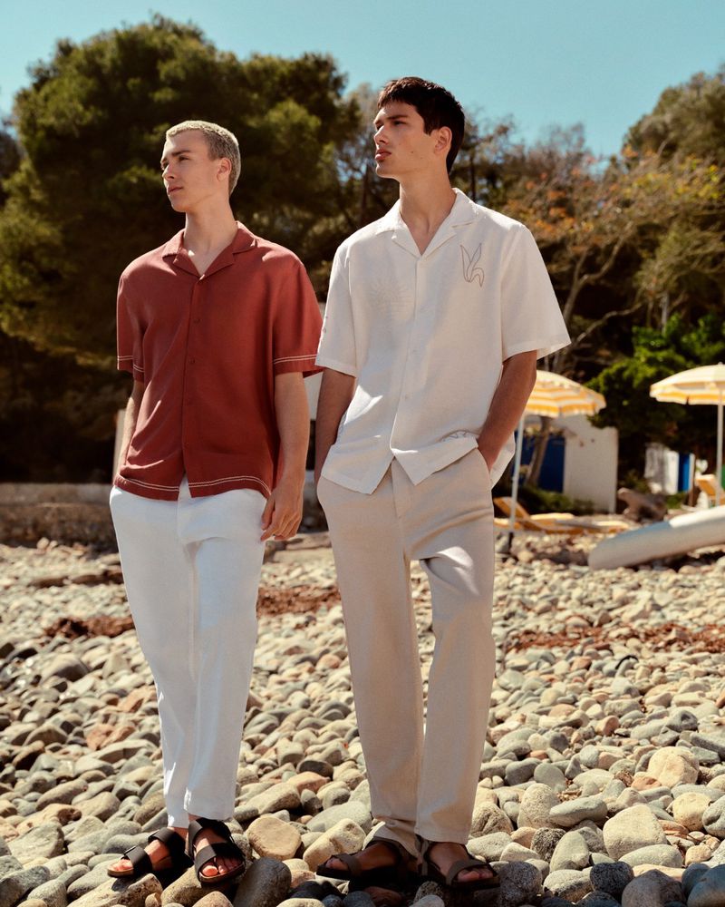 Two adult men pose outdoors on a rocky beach, wearing resort wear: button-down shirts, trousers, and sandals, near trees and beach umbrellas.