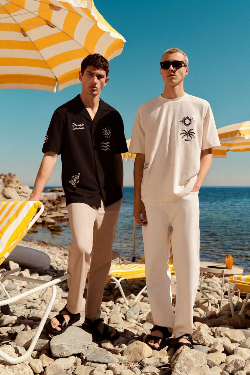 Two men model summer fashion on a rocky beach. One wears a black shirt and beige pants; the other, a white tee and pants against the blue sea.