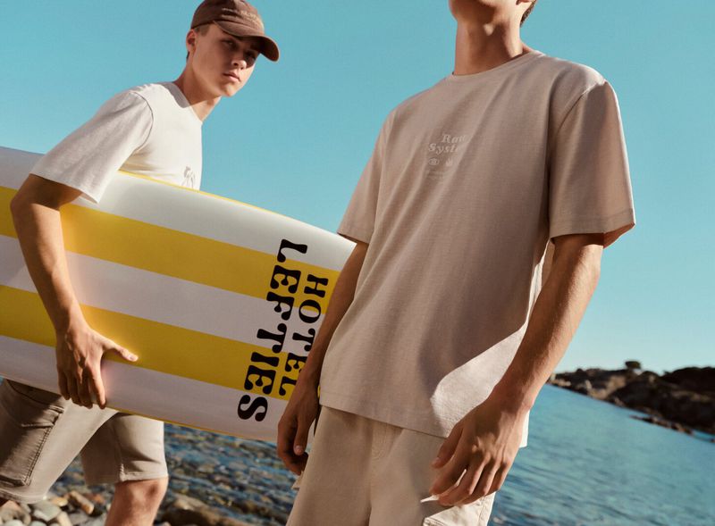Two young adult males in casual clothing stand on a rocky beach with a yellow and white surfboard against a blue sky background.