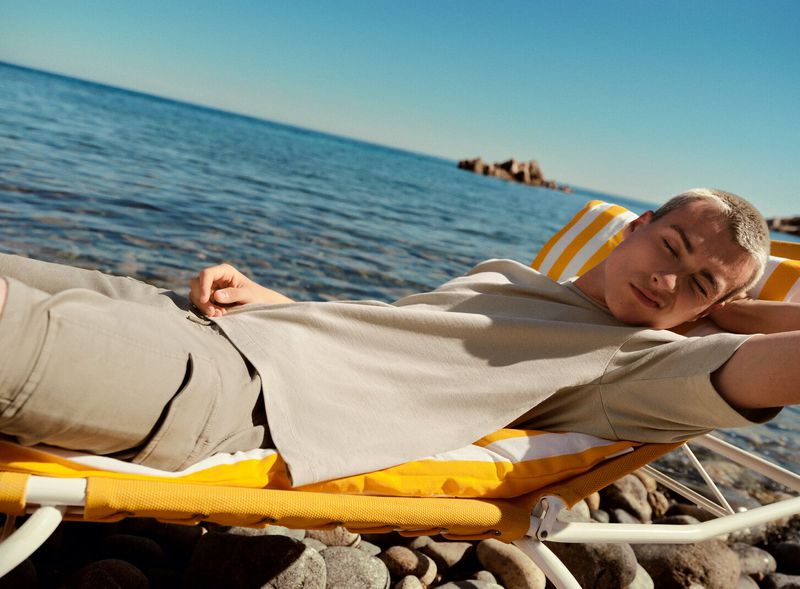 A young adult is relaxing on a yellow striped beach chair by the ocean on a sunny day with a clear blue sky in a lifestyle shot.