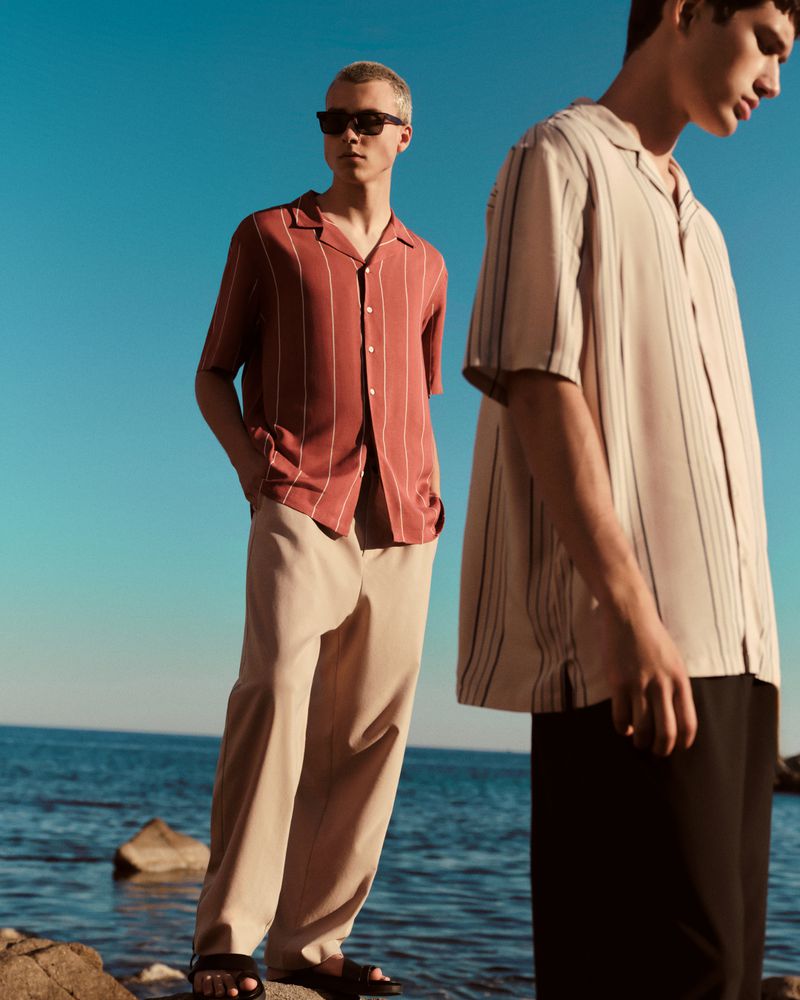 Two young adult men wearing striped shirts and casual pants pose on a rocky beach with the ocean and clear blue sky in the background.