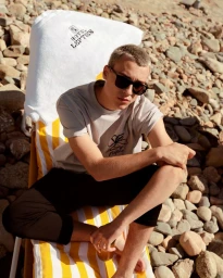 A man in sunglasses and a light t-shirt seated on a yellow striped sun lounger over a rocky beach