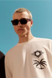 Man wearing sunglasses and a white t-shirt with a sun and palm tree graphic against a bright blue sky background.