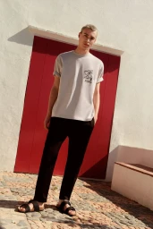 Full-length portrait of a male model wearing an off-white t-shirt and black trousers standing against a red door.
