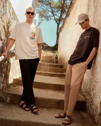 Two male models standing on stone outdoor steps wearing casual t-shirts, trousers, and sandals for Lefties Summer Collection.