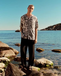 A male model standing on rocks by the ocean wearing a patterned short-sleeve shirt and black trousers for Lefties Summer Collection.