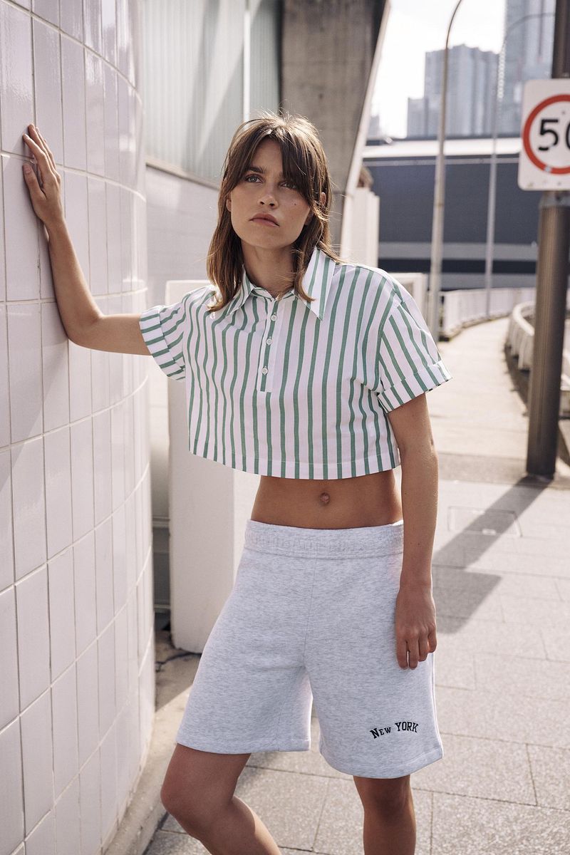 Woman in a striped crop top and athletic shorts poses outdoors against a wall, with buildings and city infrastructure in the background.