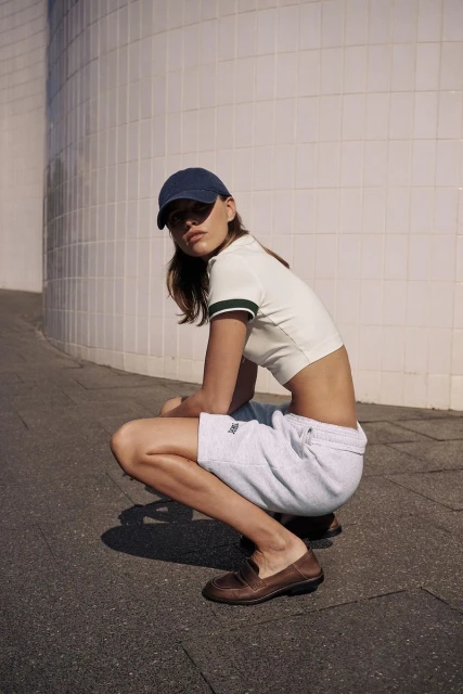 A young adult woman in athletic wear squats in dappled sunlight against a textured wall, blending fashion with casual fitness.
