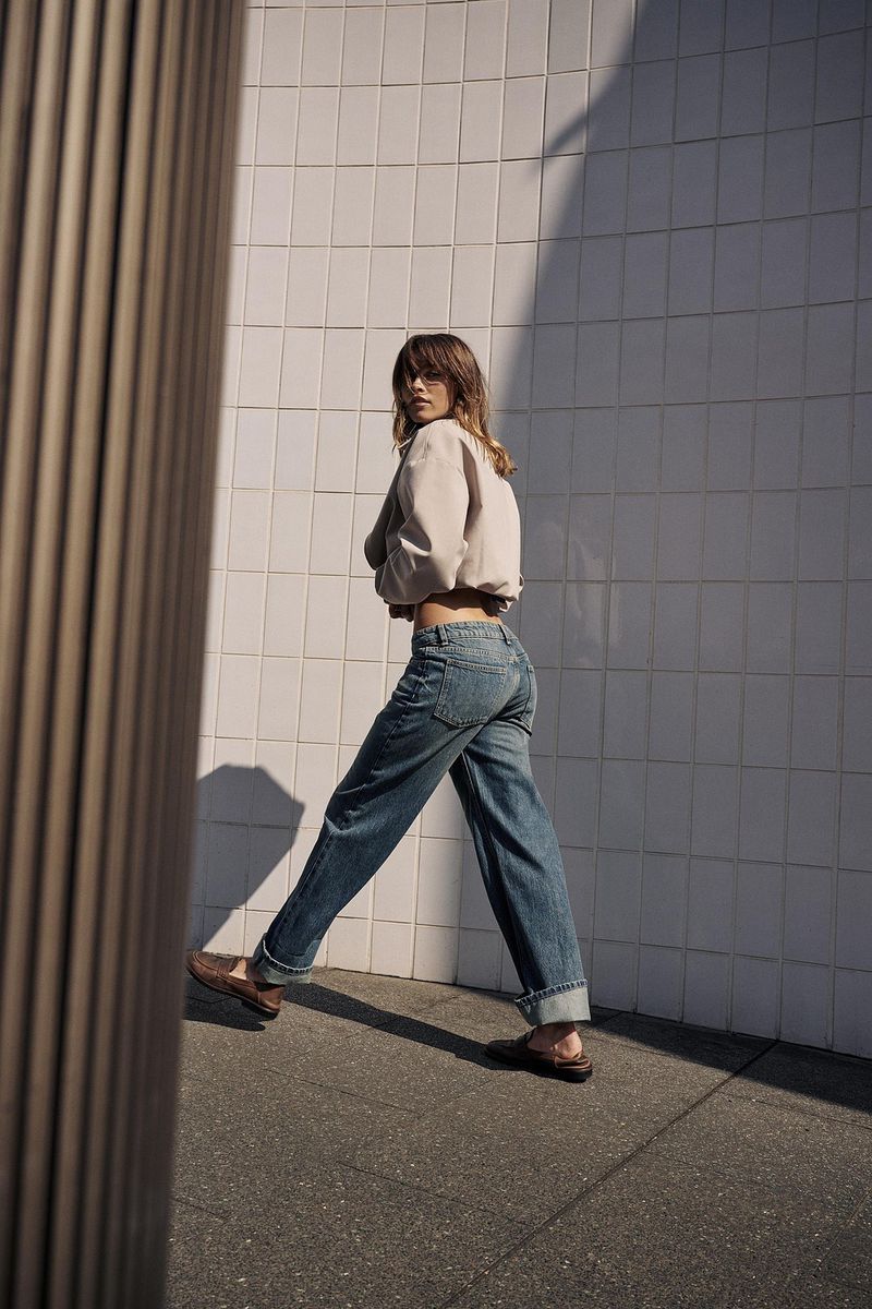 Adult woman in casual fashion turns to face the camera while walking outside in blue jeans, brown shoes, and a beige jacket.