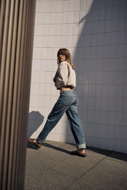 Adult woman in casual fashion turns to face the camera while walking outside in blue jeans, brown shoes, and a beige jacket.