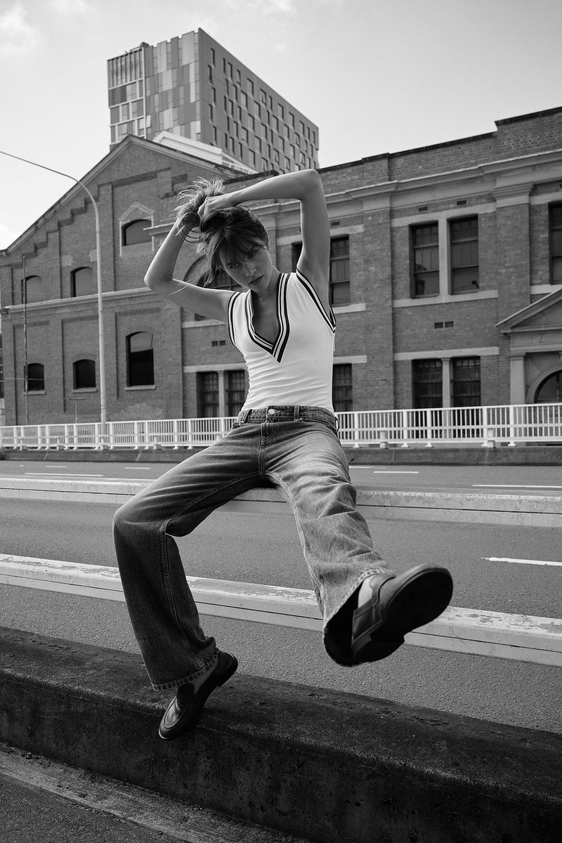 Black and white shot of a woman in jeans and a top sitting on a ledge in an urban setting, with buildings in the background.