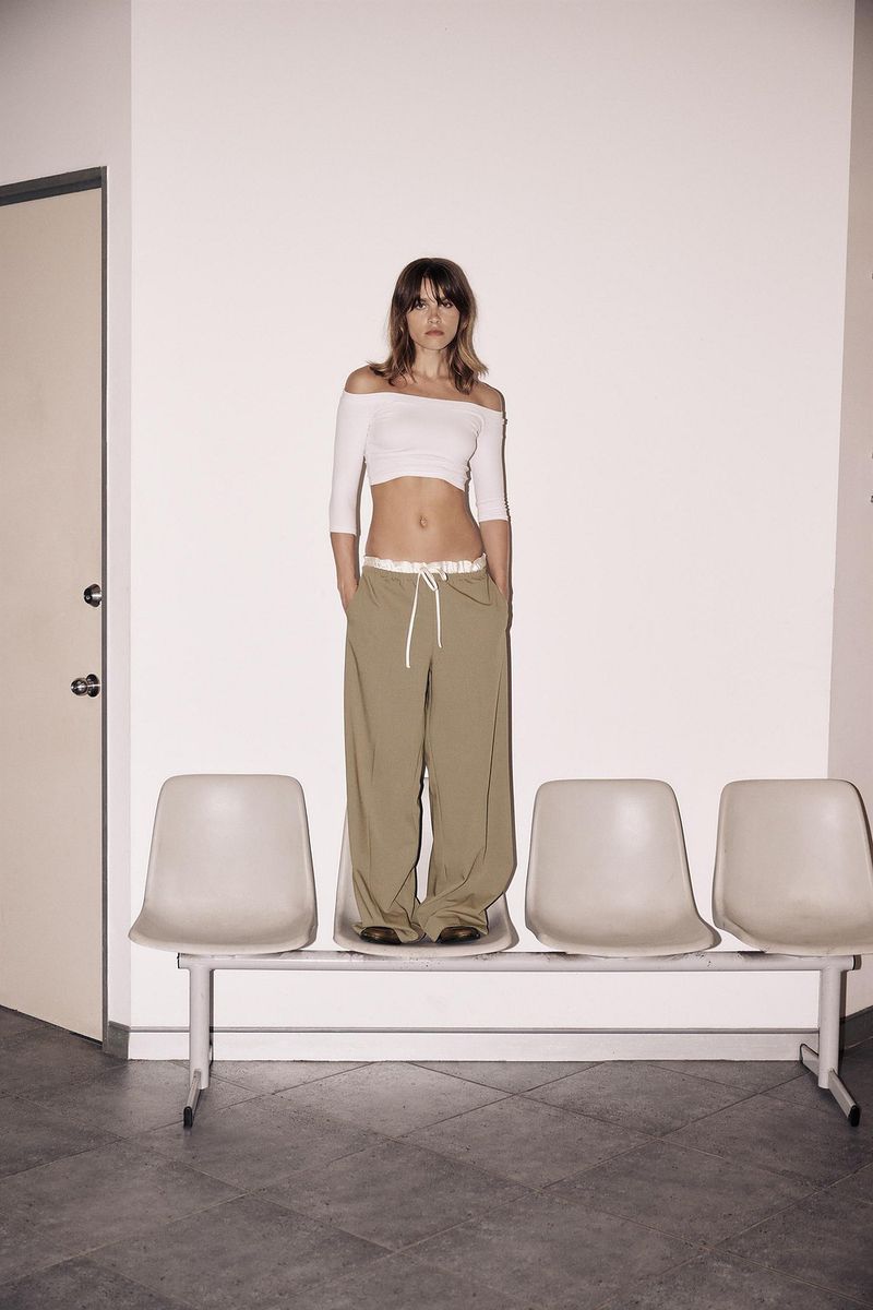 A slender woman with light brown hair stands on a bench in front of a white wall wearing an off-shoulder top and loose, neutral-colored pants.
