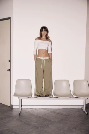 A slender woman with light brown hair stands on a bench in front of a white wall wearing an off-shoulder top and loose, neutral-colored pants.