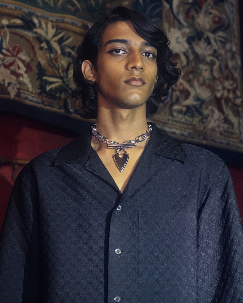 Young man with dark hair poses in front of a tapestry wearing a black button-down shirt and silver chain necklace with an arrow pendant.