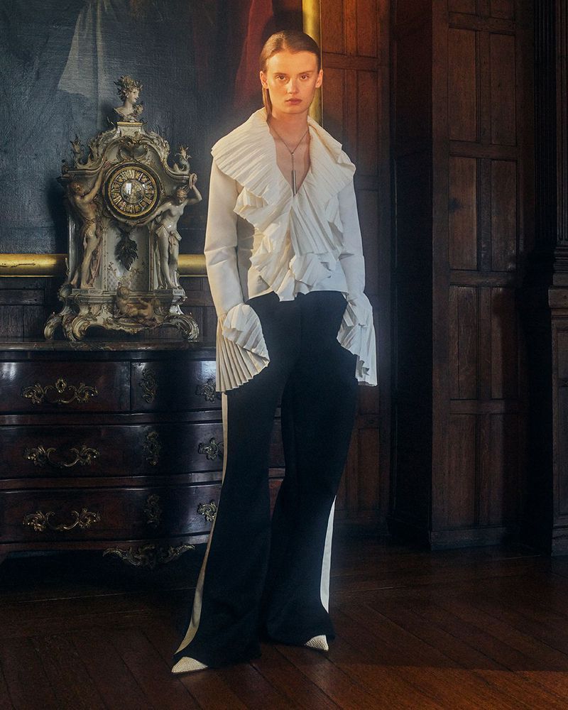 Editorial fashion shot featuring a model in an elegant white ruffled shirt and black pants in front of a dark wood wall and clock.
