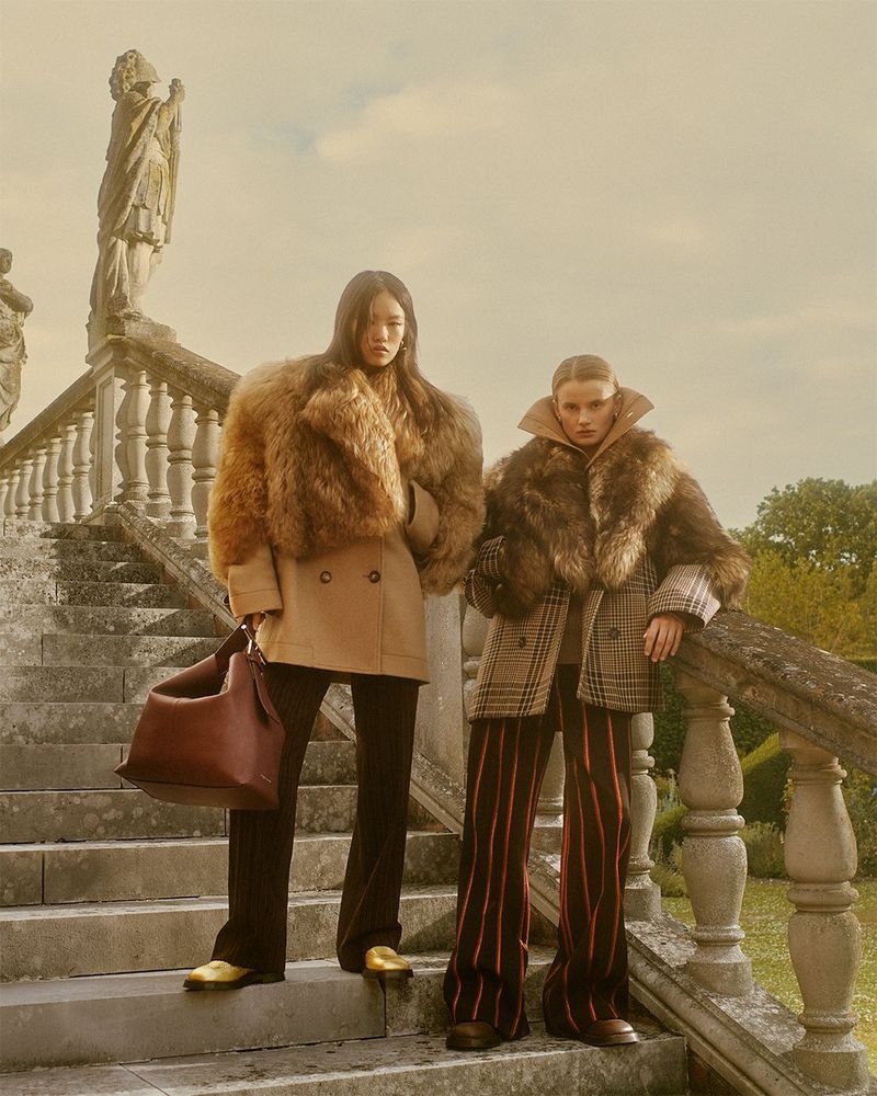 Two models on stone steps wear vintage-style coats with fur collars, dark pants, and gold shoes, standing near an old statue.