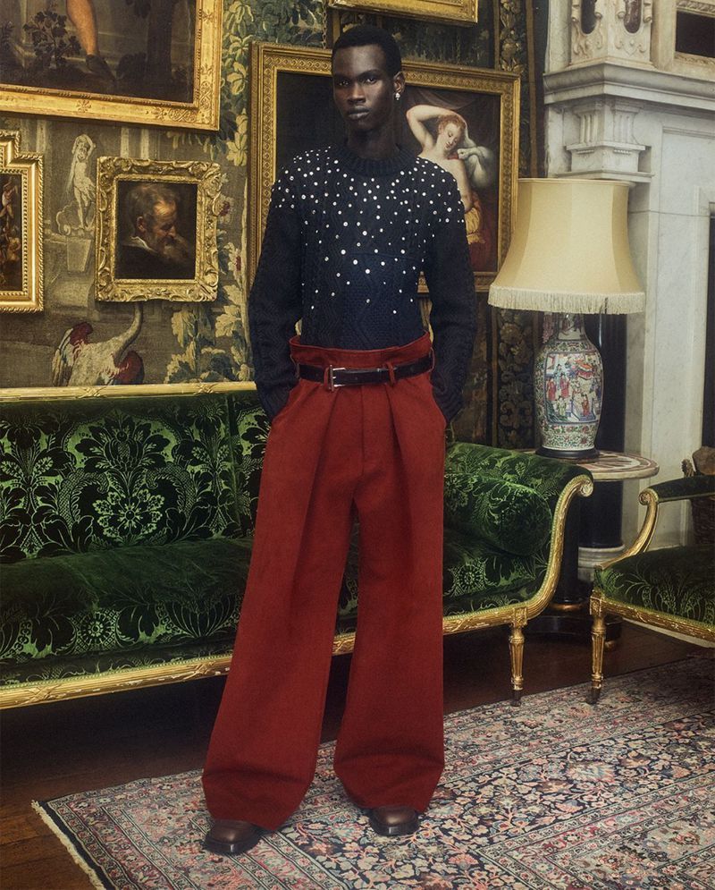 A Black male model poses in a patterned sweater and red wide-leg trousers in a traditional-style room with paintings and antique furniture.