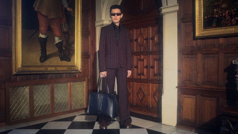 Person wearing a pinstripe suit and sunglasses holding a tote bag while standing in a wood-paneled room with paintings.