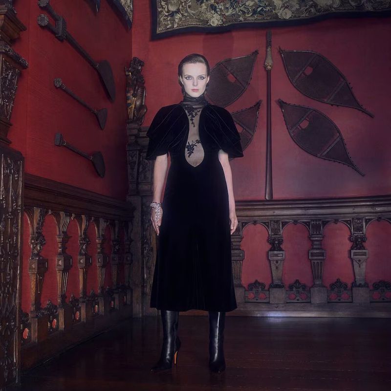 Elegant woman in black velvet gown stands in an ornate interior hall with red walls, decorative weapons, and dark wooden floors and trim.