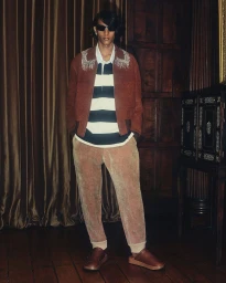 Model wearing dark sunglasses, a brown jacket, striped polo shirt, and brown trousers standing indoors.