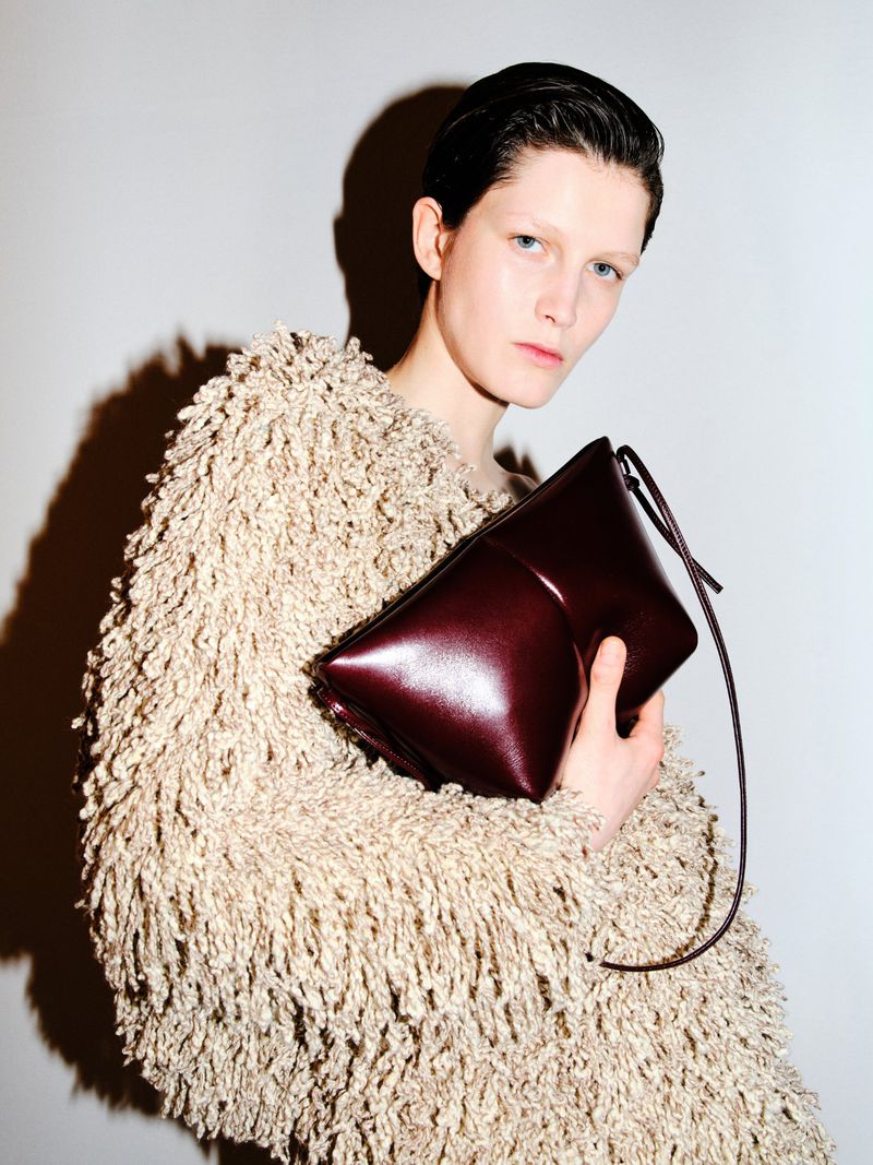 Woman poses in a shaggy tan sweater holding a structured burgundy purse against a neutral backdrop in a studio portrait fashion shoot.