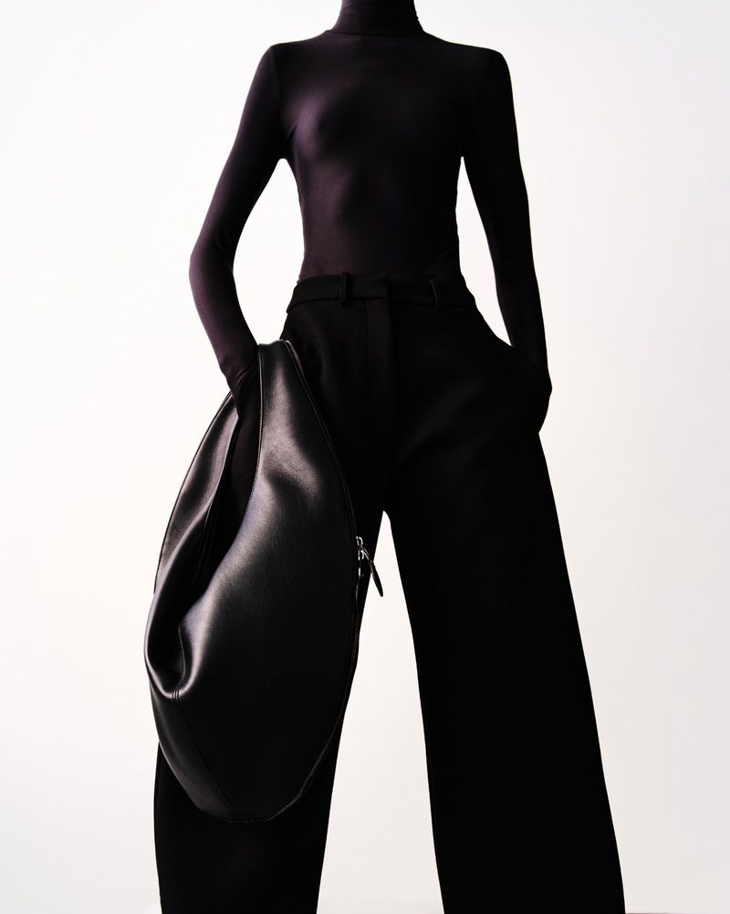 An adult fashion model poses in a black turtleneck, wide-leg pants, and a hobo bag, set against a bright white backdrop in a studio shot.