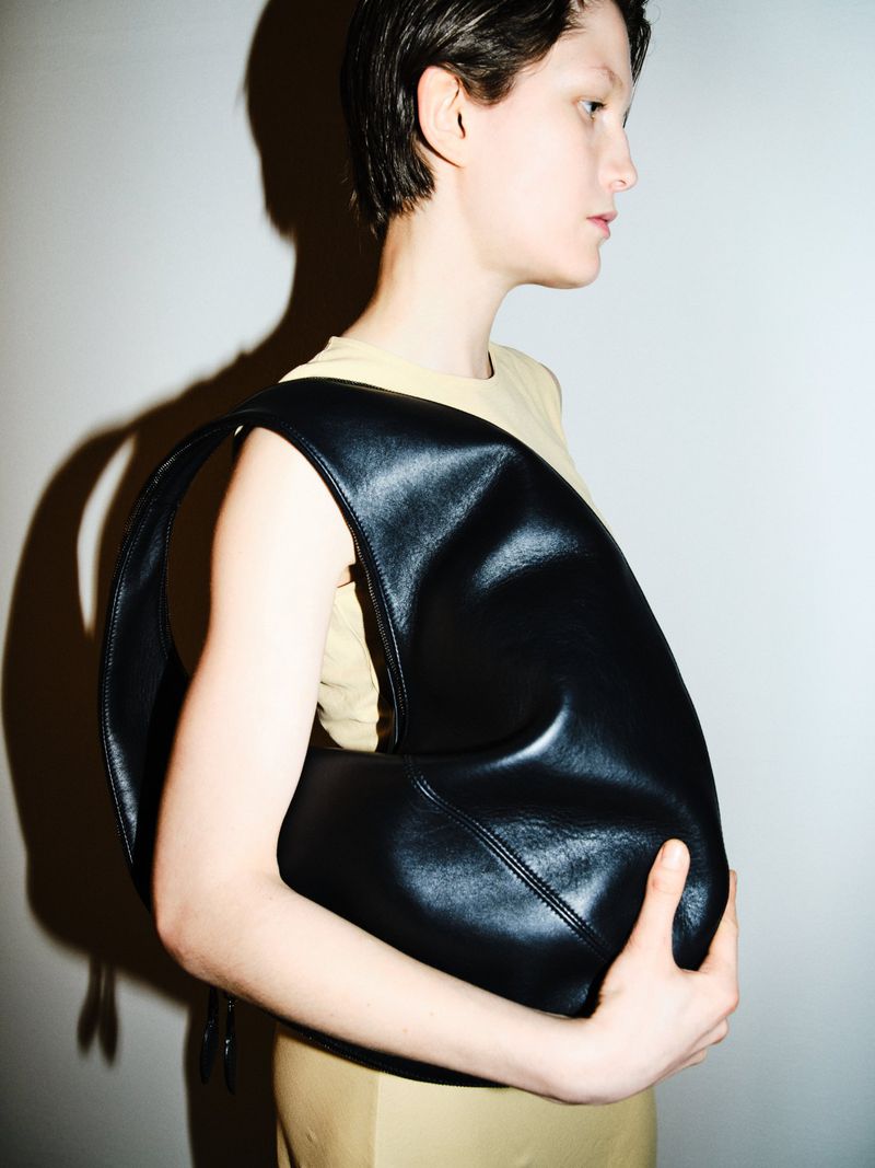 Model showcasing a black leather hobo bag against a neutral background. Fashion shot with soft, diffused lighting and a modern aesthetic.