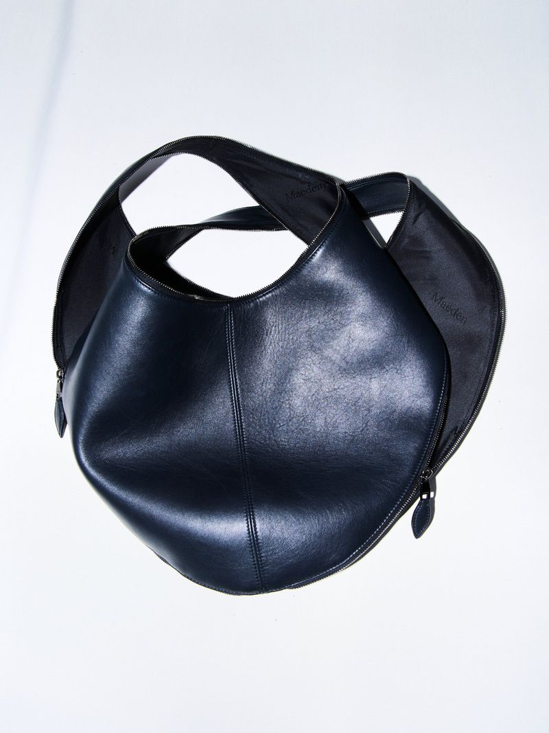 High-angle studio shot showcases a black leather hobo bag with a zip closure, featuring a modern design and sleek craftsmanship against white.